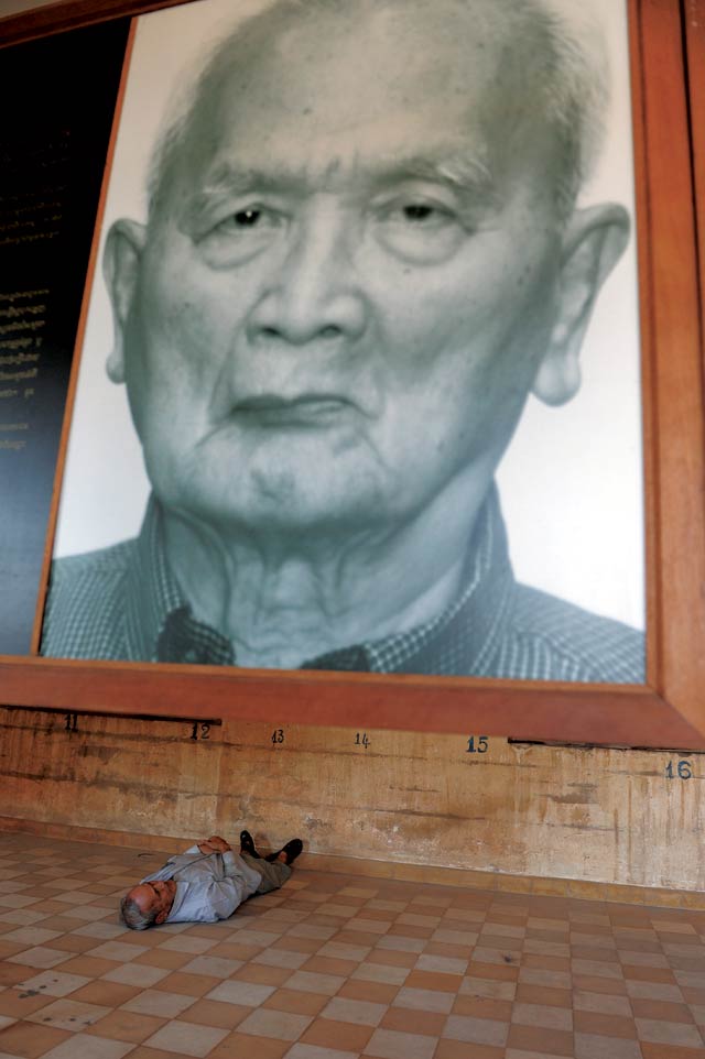 Photos: Tang Chhin Sothy, ECCC. Hidden truth: Tuol Sleng survivor Bou Meng displays the manner in which he was once shackled, in front of a portrait of former Khmer Rouge leader Nuon Chea.